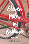 Eleven Miles to Oshkosh: Cover art of a close up picture of a red bicycle's back tire. The bike is on a dirt road. The title in written in a thick, curly, white font.
