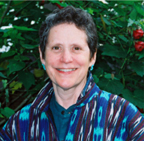 This is a photo of the author, Susan Krieger. She is wearing a blue, aqua and purple pattern sweater over a green shirt. On the leafy background behind her, two red flowers can be seen. She is wearing aqua earrings.