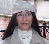 the author, Paola Corso, leans against a brick wall, she is wearing a light colored béret.